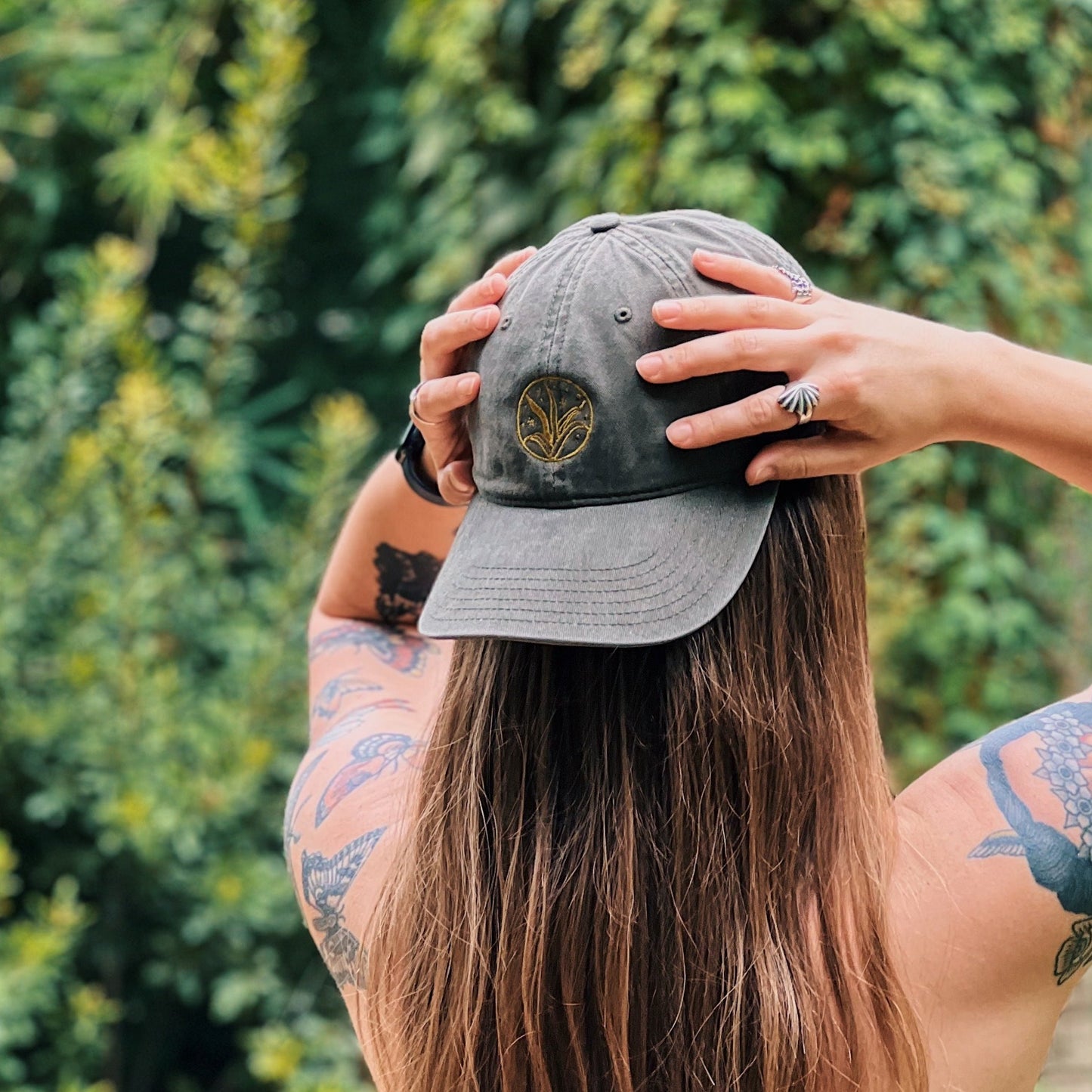Photograph of the backside of person wearing an olive green 'dad hat' with light green embroidery in the shape of an agave plant, backward on their head with greenery out of focus in the background