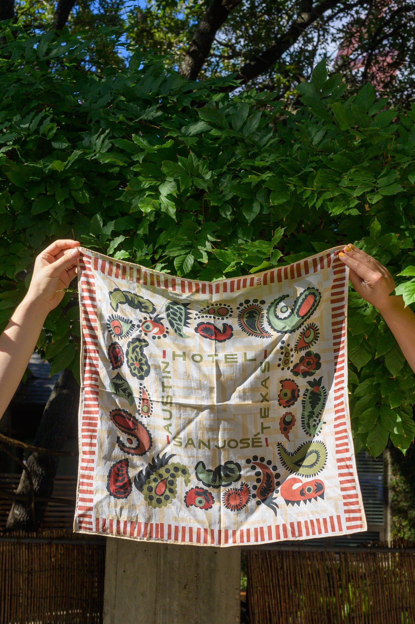 A bandana with a white and cream base featuring red and green paisley patterns and text that reads 'Austin', 'Hotel San Jose', and 'Texas' being held up by two hands in front of greenery on an outside lounge.