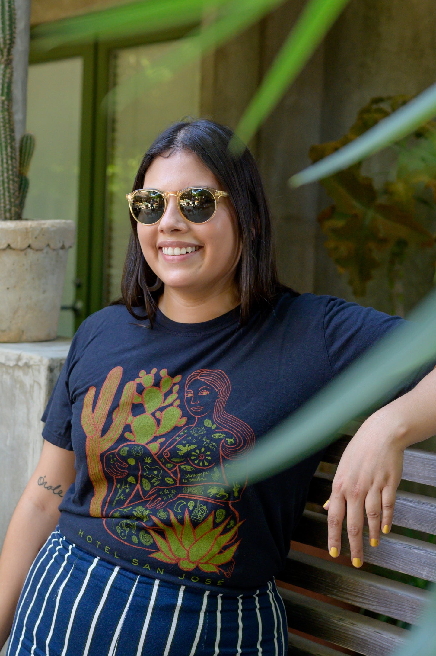 Woman wearing a navy tshirt with line work illustration of a woman covered in Austin Texas icons with various cacti in front and behind her with text that reads 'HOTEL SAN JOSE' underneath. , standing outdoors.