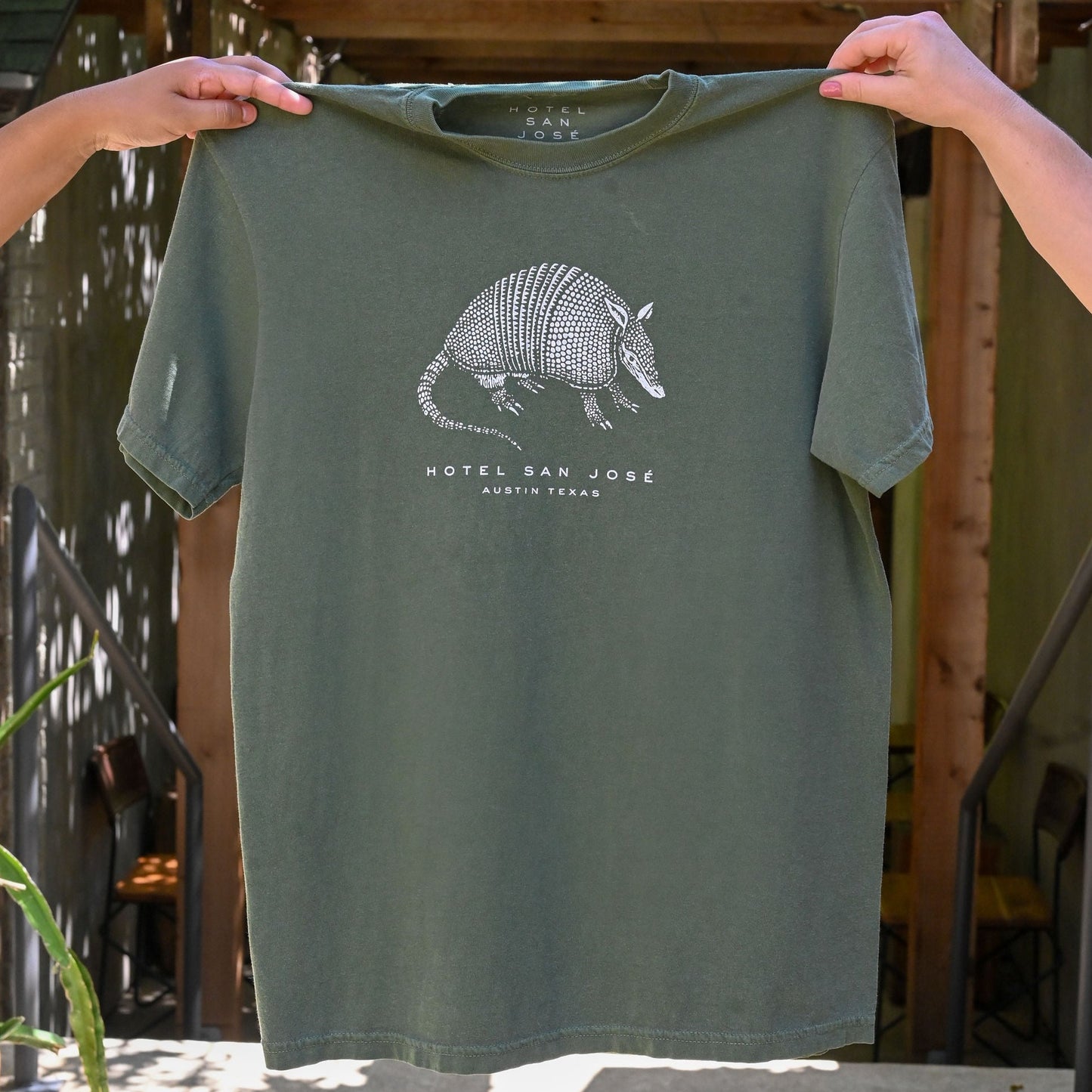 A green unisex crew neck t-shirt with a white printed armadillo design and text 'HOTEL SAN JOSE AUSTIN TEXAS' on the front being held by hands in front of a green brick and wooden structure