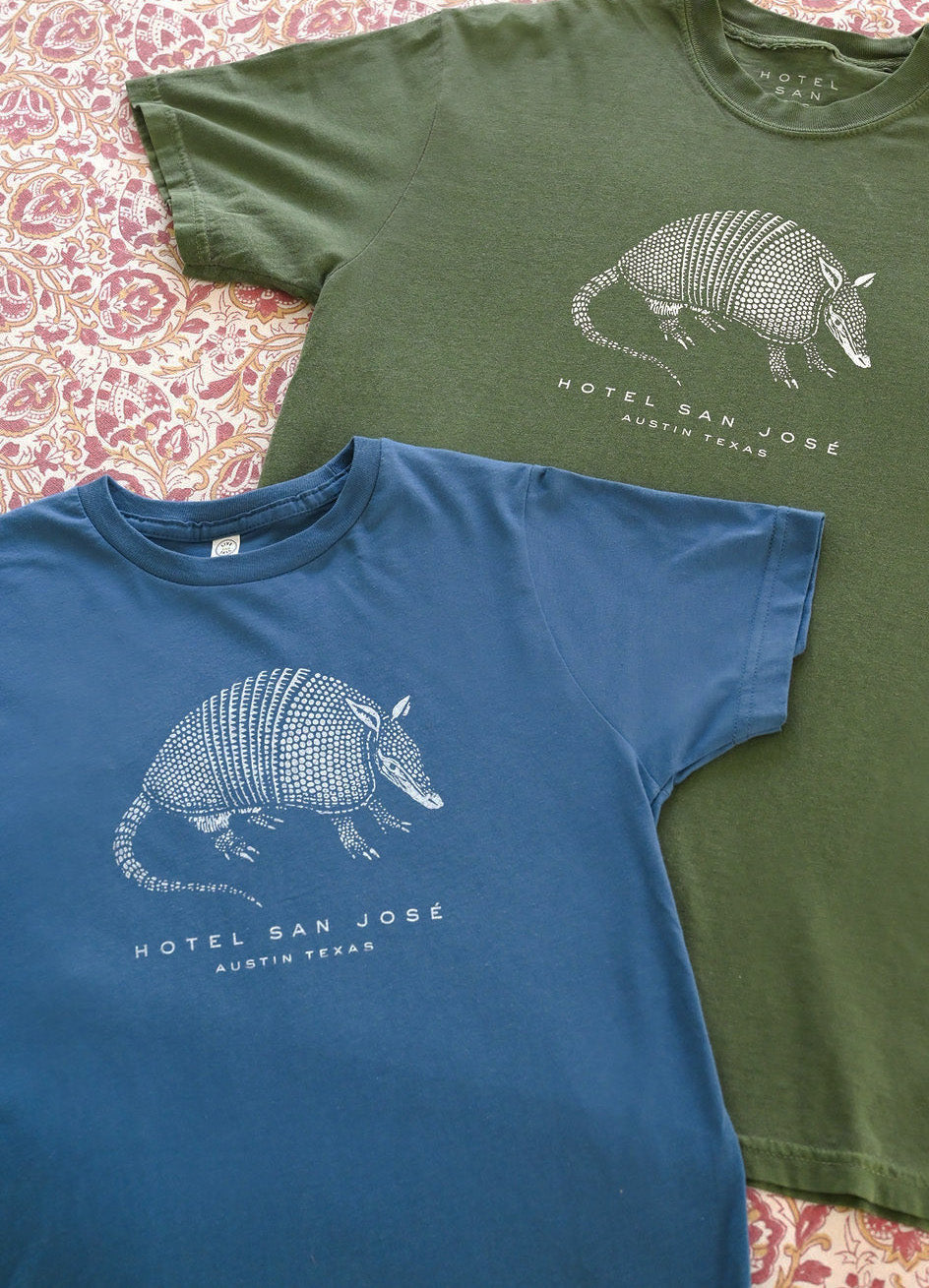 Two t-shirts, one green and one blue, with an armadillo graphic and text 'HOTEL SAN JOSE AUSTIN TEXAS' on a patterned surface.