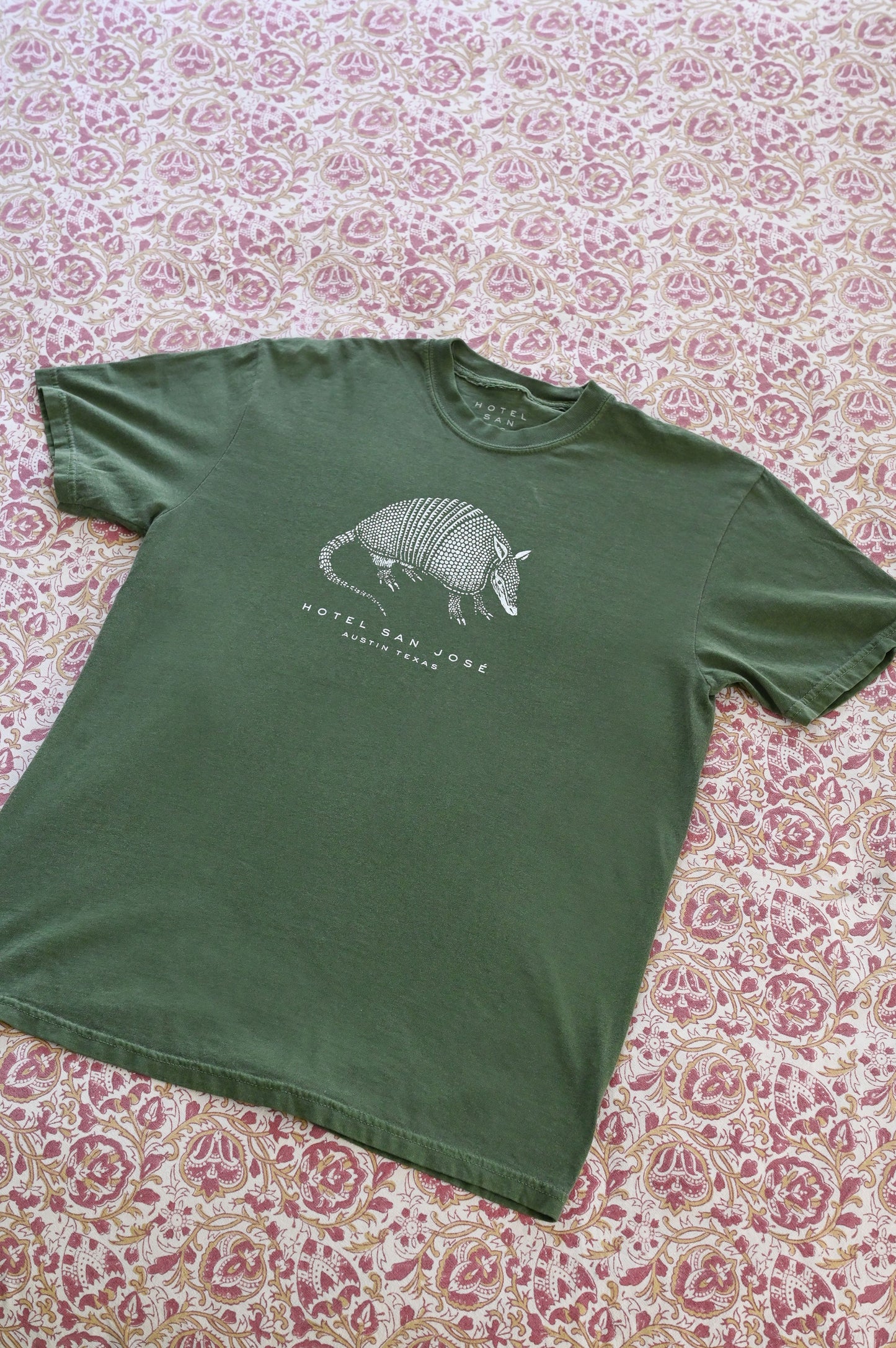 A green unisex crew neck t-shirt with a white printed armadillo design and text 'HOTEL SAN JOSE AUSTIN TEXAS' on the front laying on a floral patterned textile.