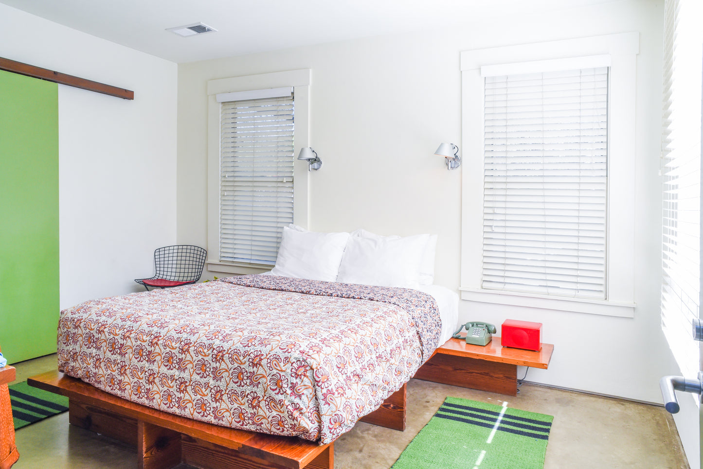 Hotel room with a bed with floral duvet cover on top, nightstands, green door and green small rugs and chairs near windows