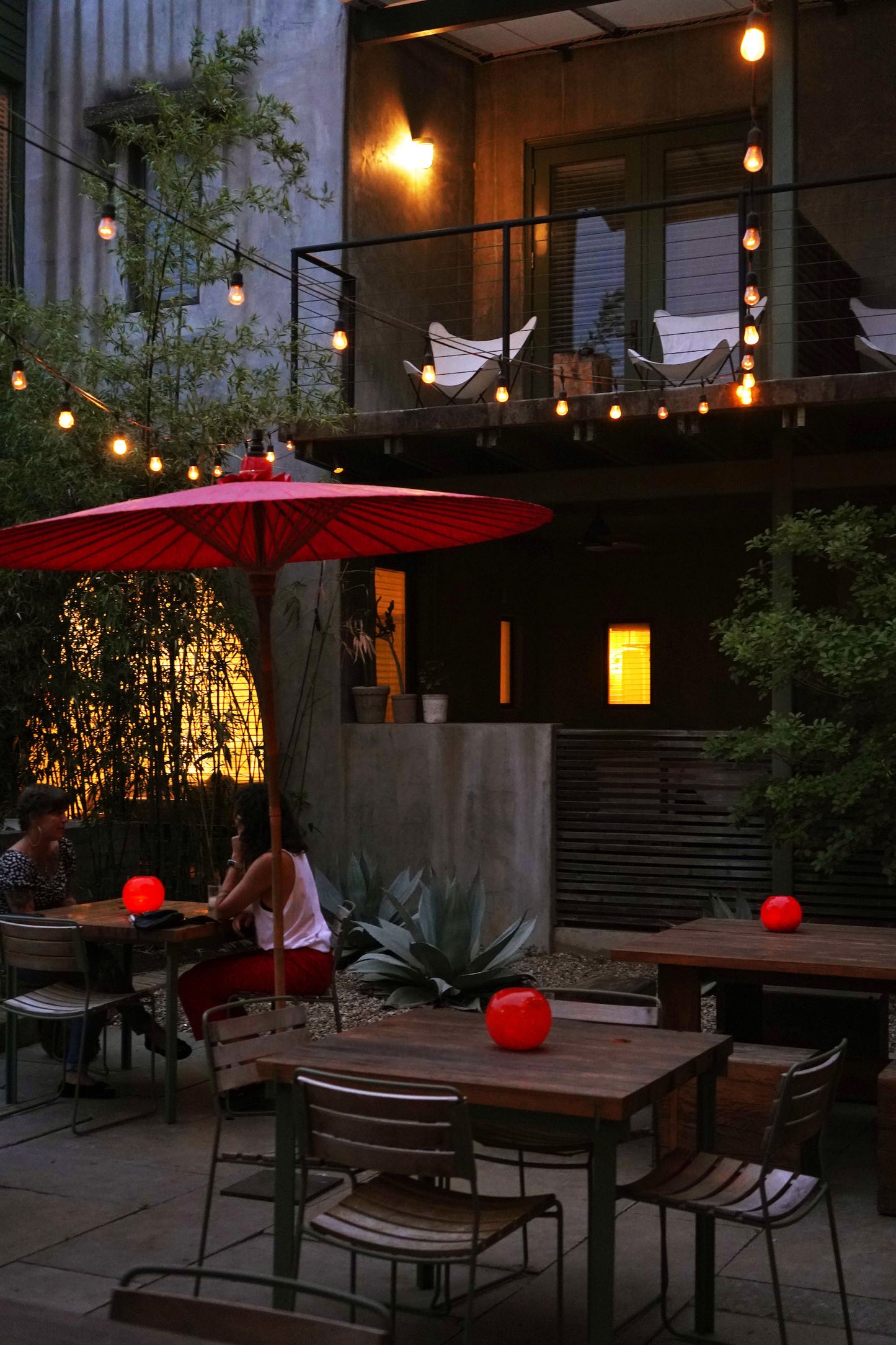 Moody photo of hotel san jose outside lounge with red glowing glass globes sitting on top of wooden and metal table sets with hotel room balcony in background.
