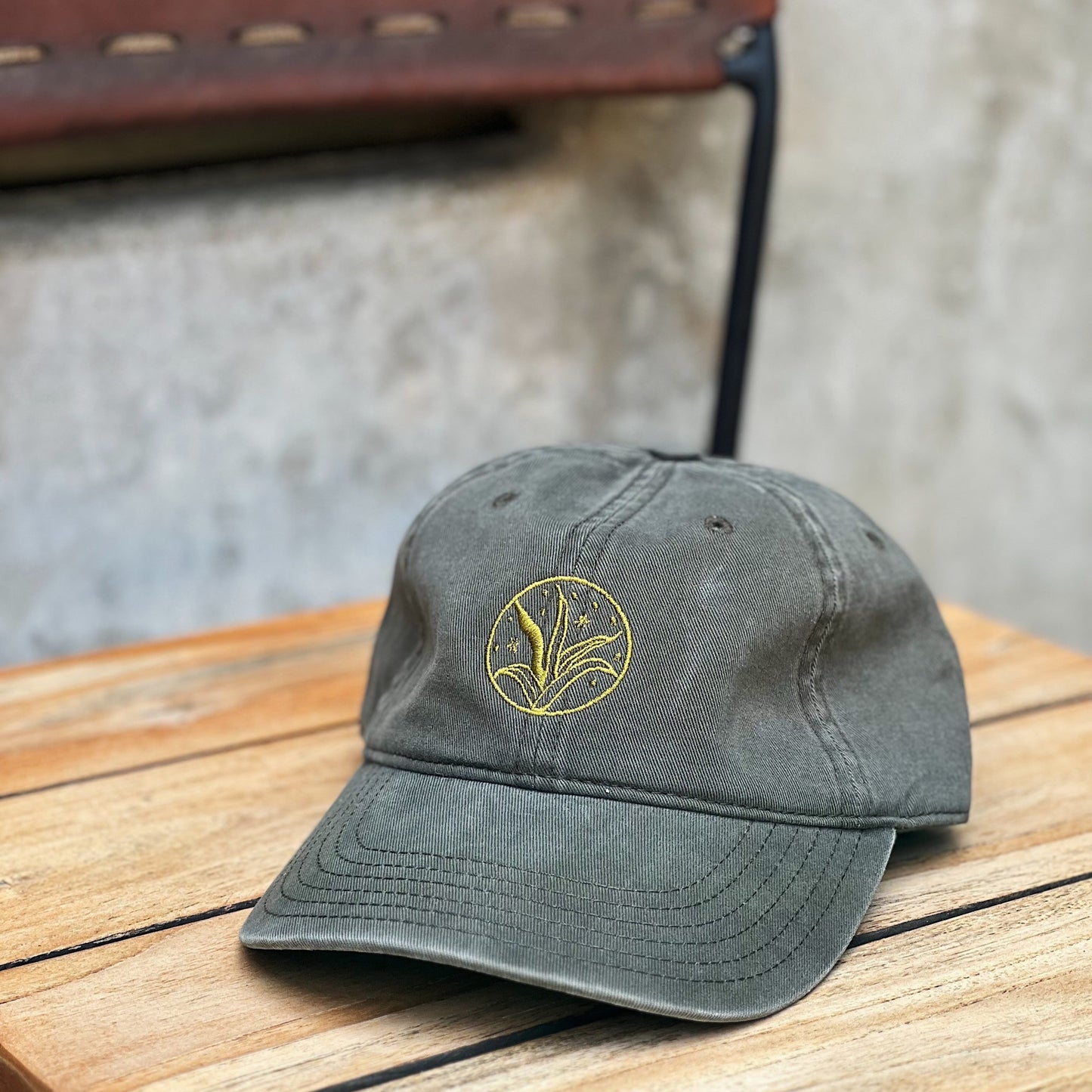 An olive green 'dad hat' with light green embroidery in the shape of an agave plant sitting on top of wooden and leather chair with cement in the background.