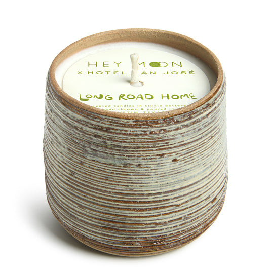 A cylindrical ceramic candle vessel with a striped pattern, including paper topper branded with 'Hey Moon x Hotel San Jose' and the name 'Long Road Home'.