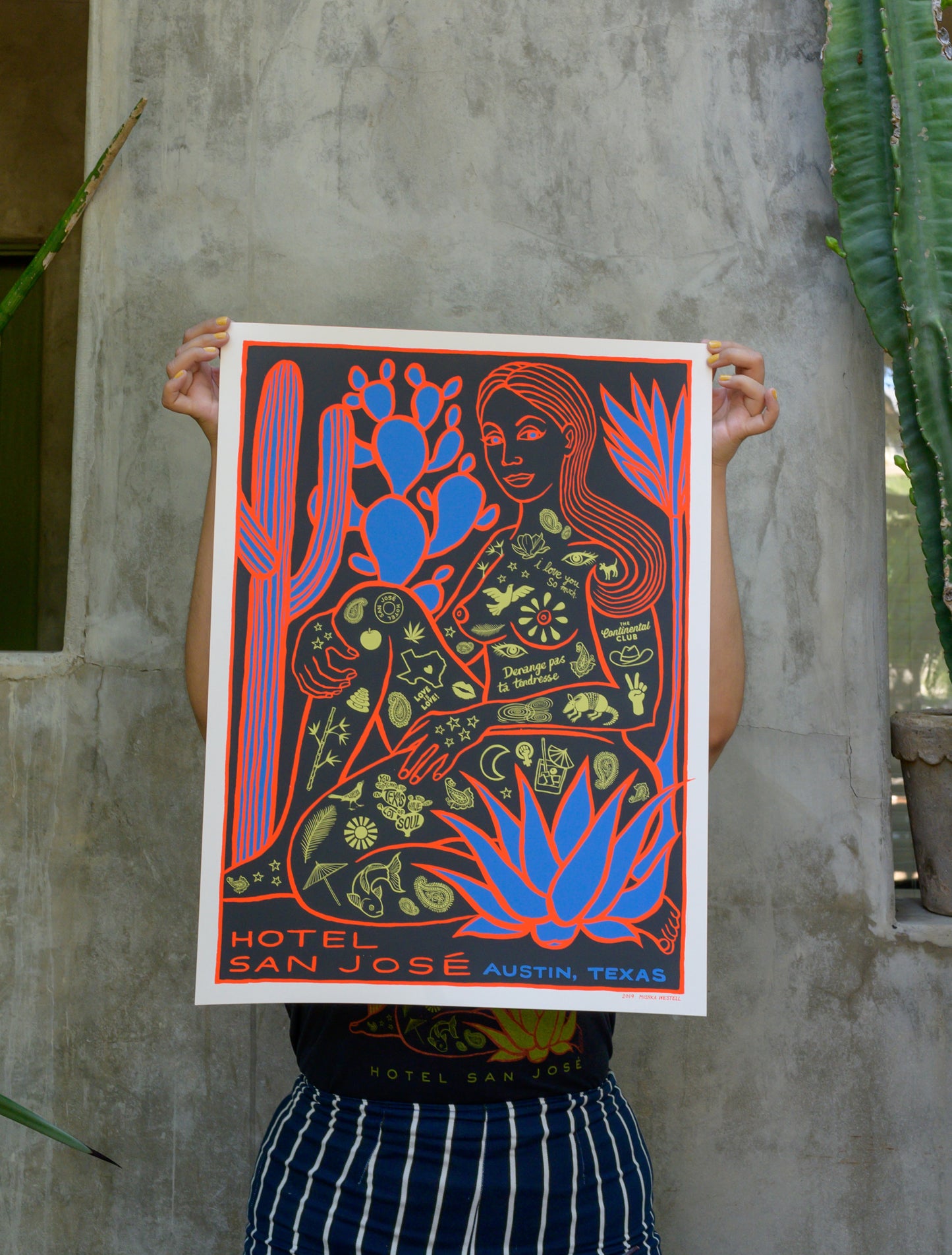 A poster featuring a stylized illustration of a woman covered in Austin's icon tattoos, sitting among cacti, with the text 'Hotel San Jose Austin, Texas' at the bottom being held by a person in front of cement wall with cacti plants on either side.