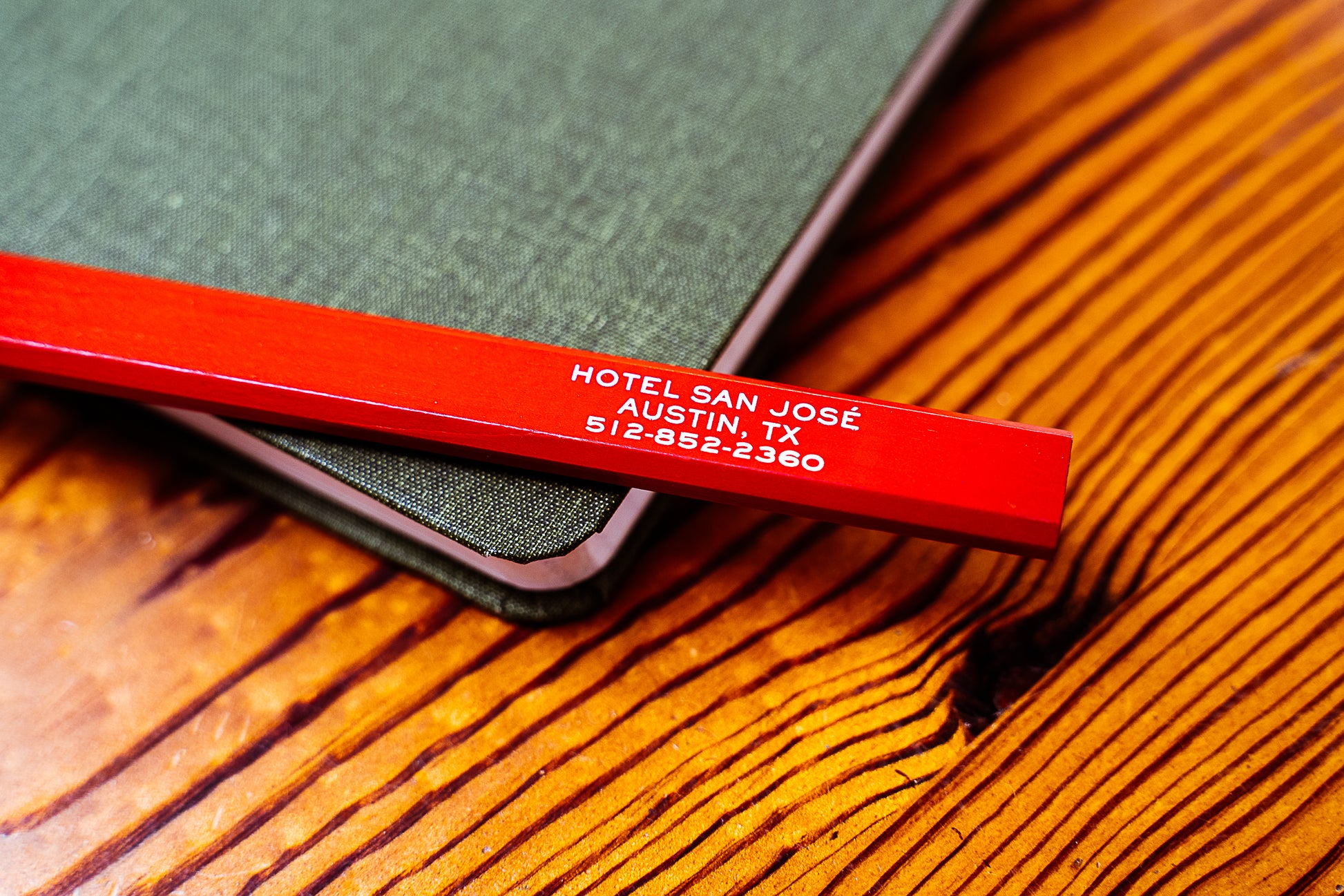 A red carpenter pencil with 'Hotel San Jose' printed in white text on the side sitting atop a notebook on a wooden surface