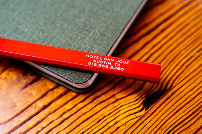 A red carpenter pencil with 'Hotel San Jose' printed in white text on the side sitting atop a notebook on a wooden surface