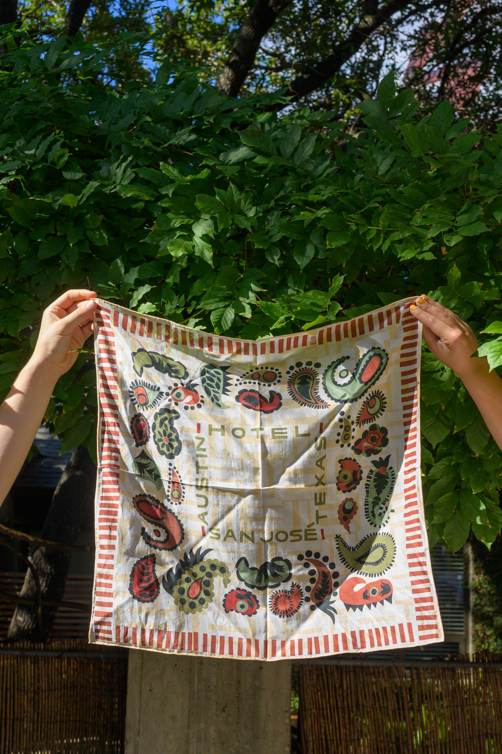 A bandana with a white and cream base featuring red and green paisley patterns and text that reads 'Austin', 'Hotel San Jose', and 'Texas' being held up by two hands in front of greenery on an outside lounge.