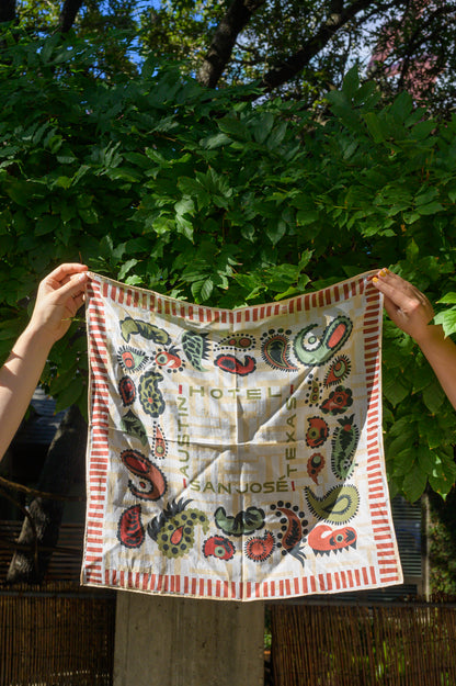 A bandana with a white and cream base featuring red and green paisley patterns and text that reads 'Austin', 'Hotel San Jose', and 'Texas' being held up by two hands in front of greenery on an outside lounge.