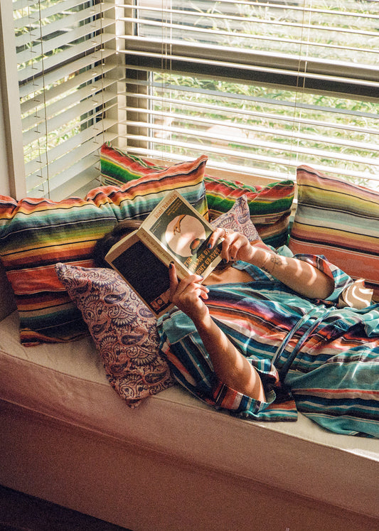 Person wearing striped serape blue robe laying on a window bed with other similar striped pillows around, reading a book with their face covered.