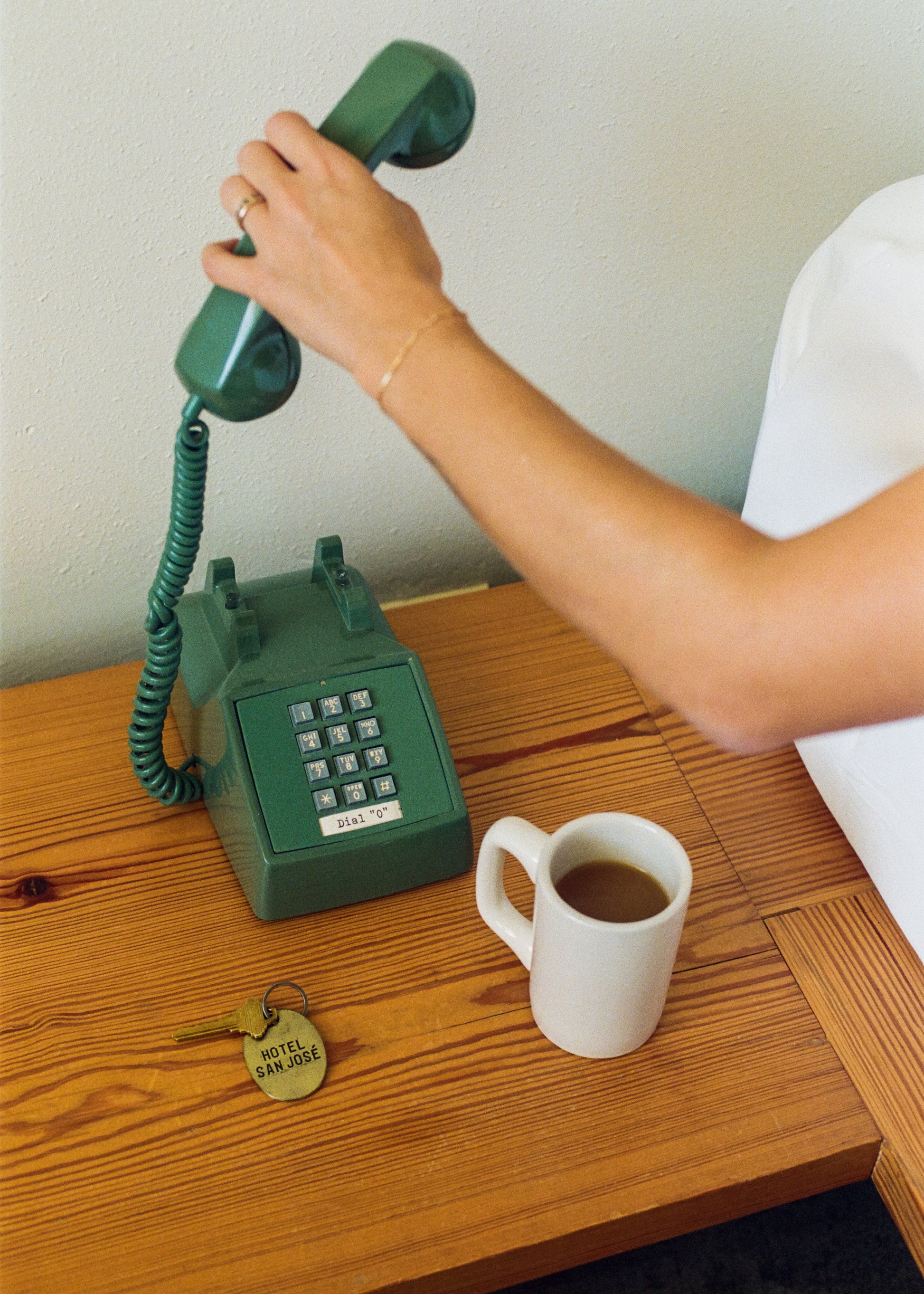 Photograph of arm holding vintage green telephone on wooden table with coffee cup and brass hotel keychain.