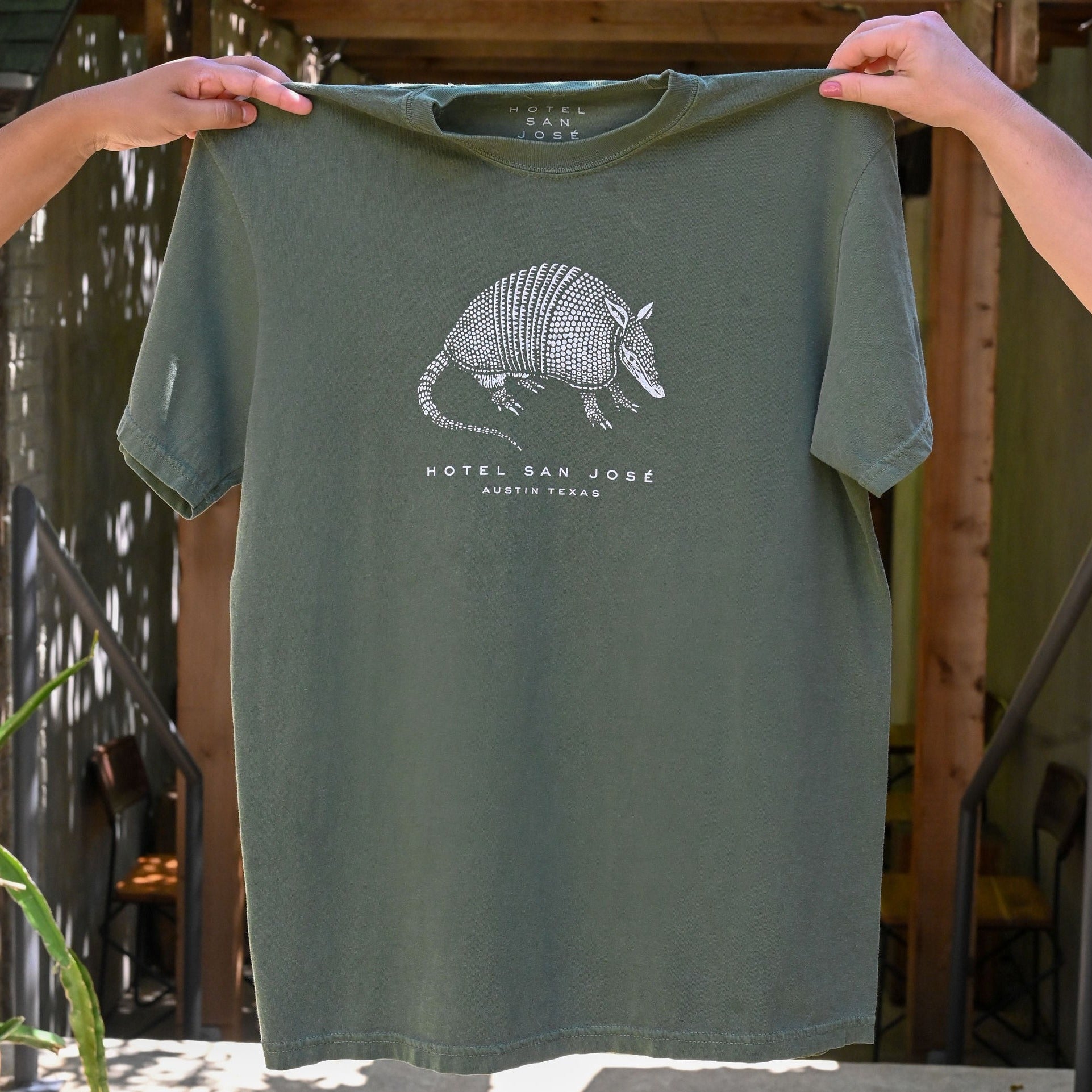 A green unisex crew neck t-shirt with a white printed armadillo design and text 'HOTEL SAN JOSE AUSTIN TEXAS' on the front being held by hands in front of a green brick and wooden structure