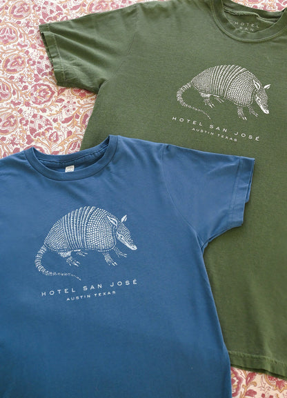 Two t-shirts, one green and one blue, with an armadillo graphic and text 'HOTEL SAN JOSE AUSTIN TEXAS' on a patterned surface.