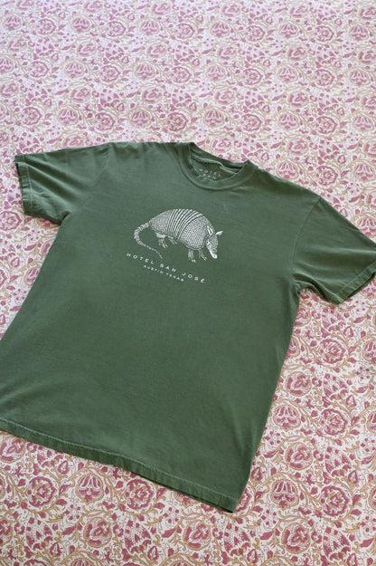 A green unisex crew neck t-shirt with a white printed armadillo design and text 'HOTEL SAN JOSE AUSTIN TEXAS' on the front laying on a floral patterned textile.
