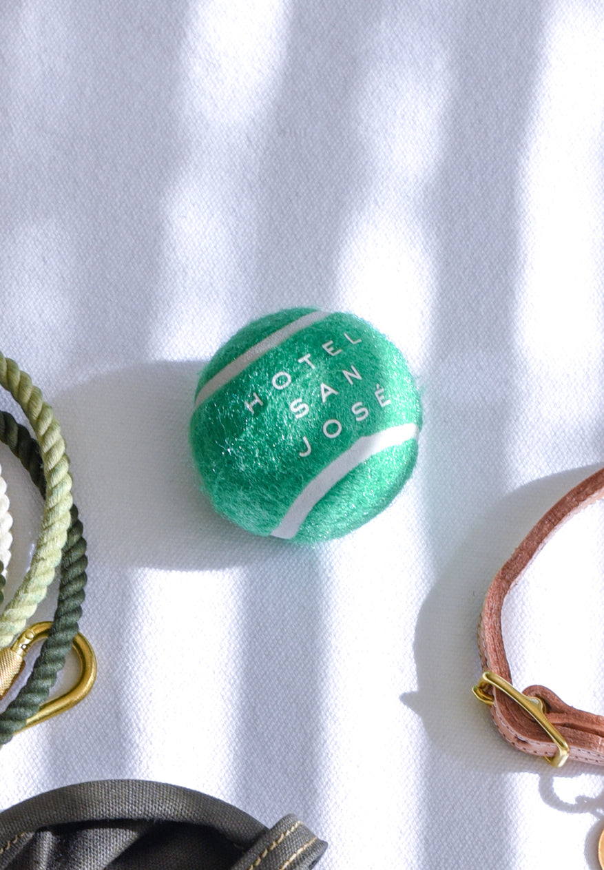 Green tennis ball with 'Hotel San Jose' text on a white surface with other dog accessories in the periphery 