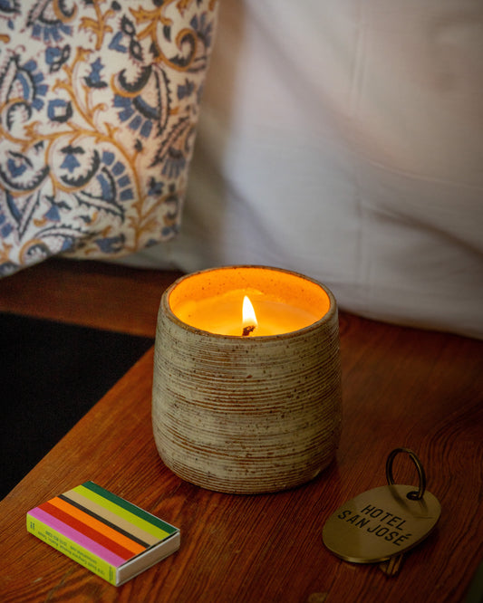 Ceramic candle in a textured holder with a lit flame on a wooden surface, next to colorful matchbox and a Hotel San Jose key tag.