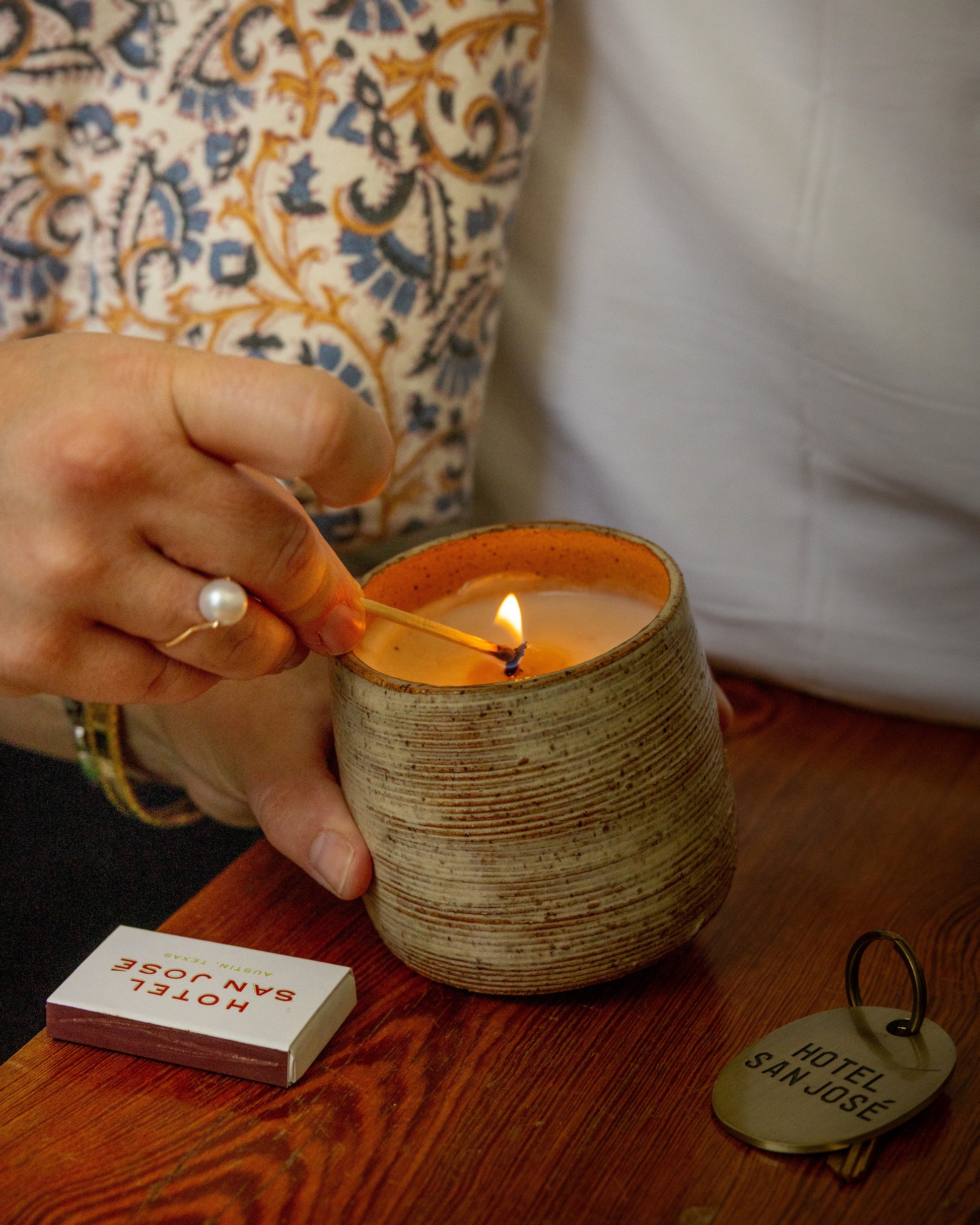 Person lighting a candle in a textured Ceramic vessel with a lit flame on a wooden surface, next to colorful matchbox and a Hotel San Jose key tag.