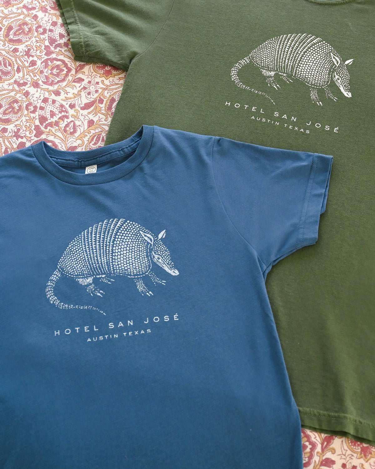 Two t-shirts, one blue and one green, with armadillo graphics and text on a patterned surface.