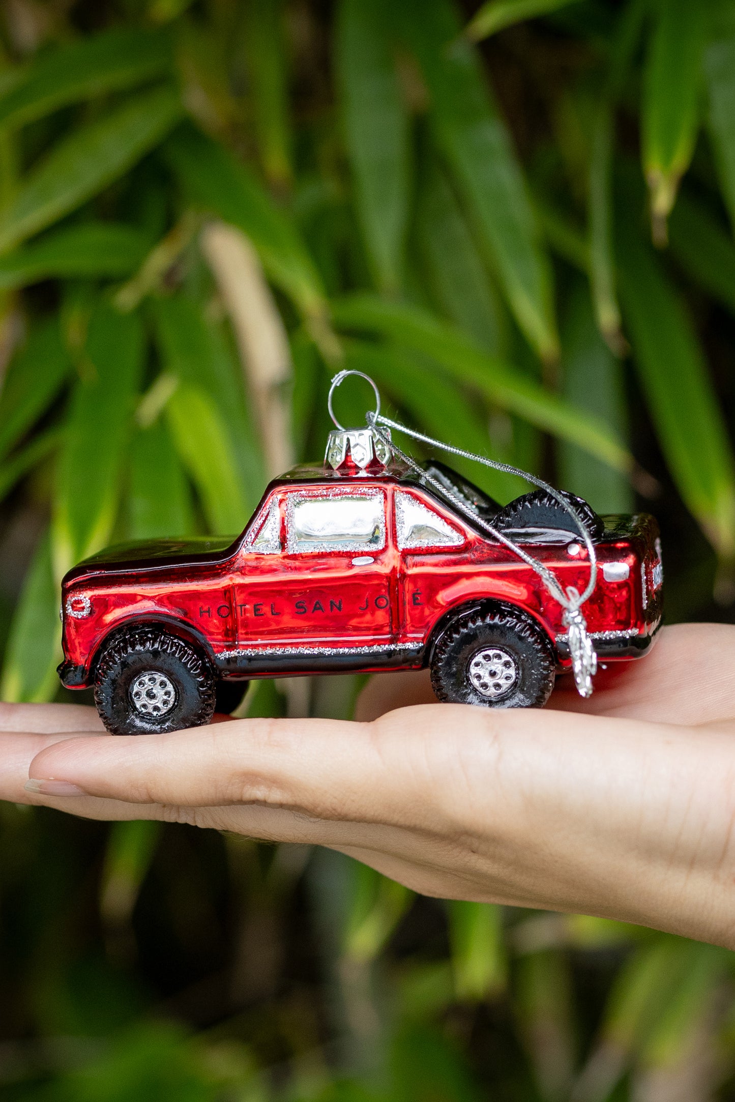 Christmas tree ornament shaped like Red truck with text 'HOTEL SAN JOSE' on the side of it being held by a hand with greenery as background.