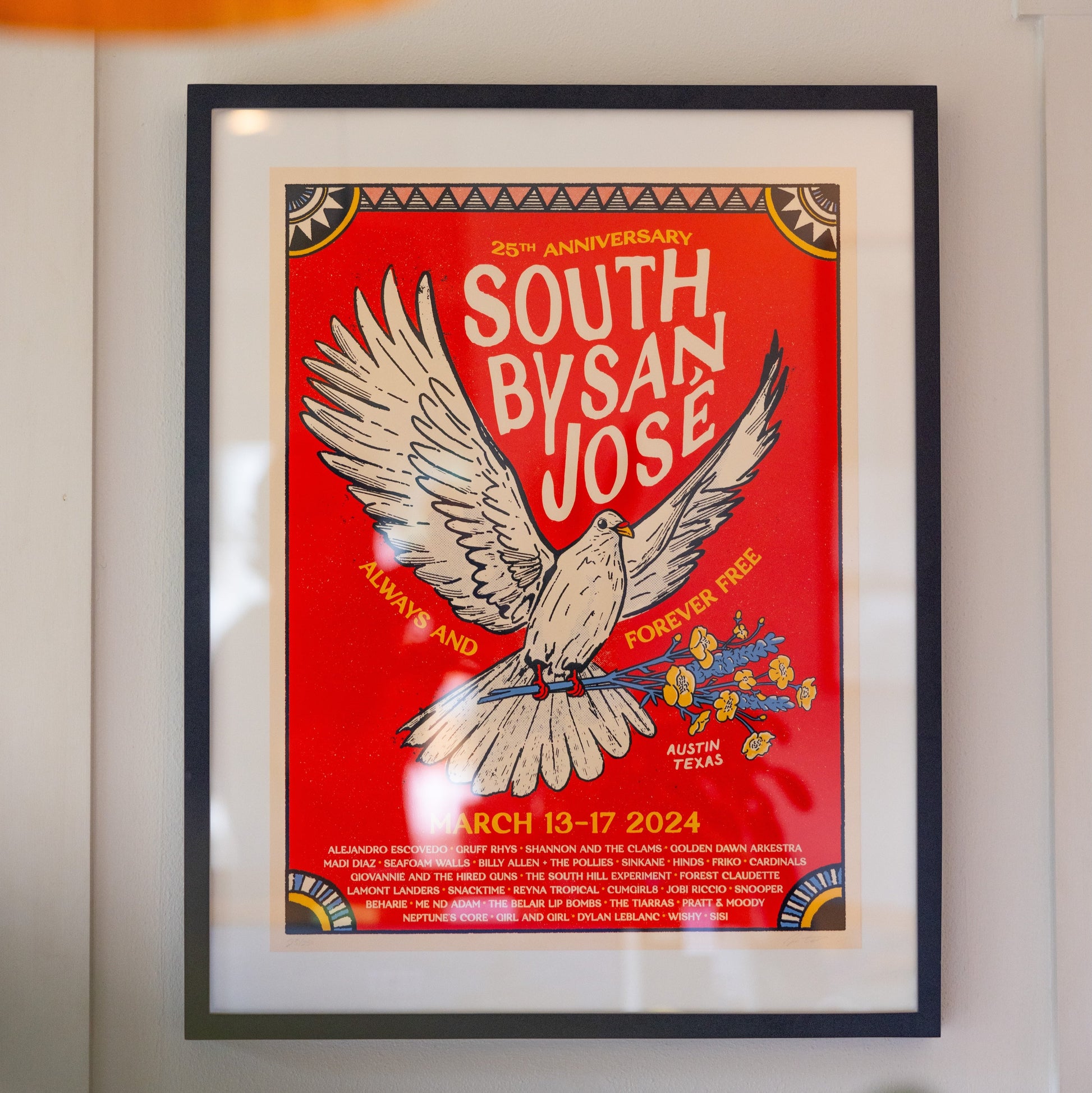 A colorful poster featuring a bird with spread wings, text celebrating the 25th Anniversary of South By San Jose, and a list of musicians in a frame, hanging on a white wall.