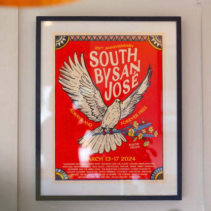 A colorful poster featuring a bird with spread wings, text celebrating the 25th Anniversary of South By San Jose, and a list of musicians in a frame, hanging on a white wall.