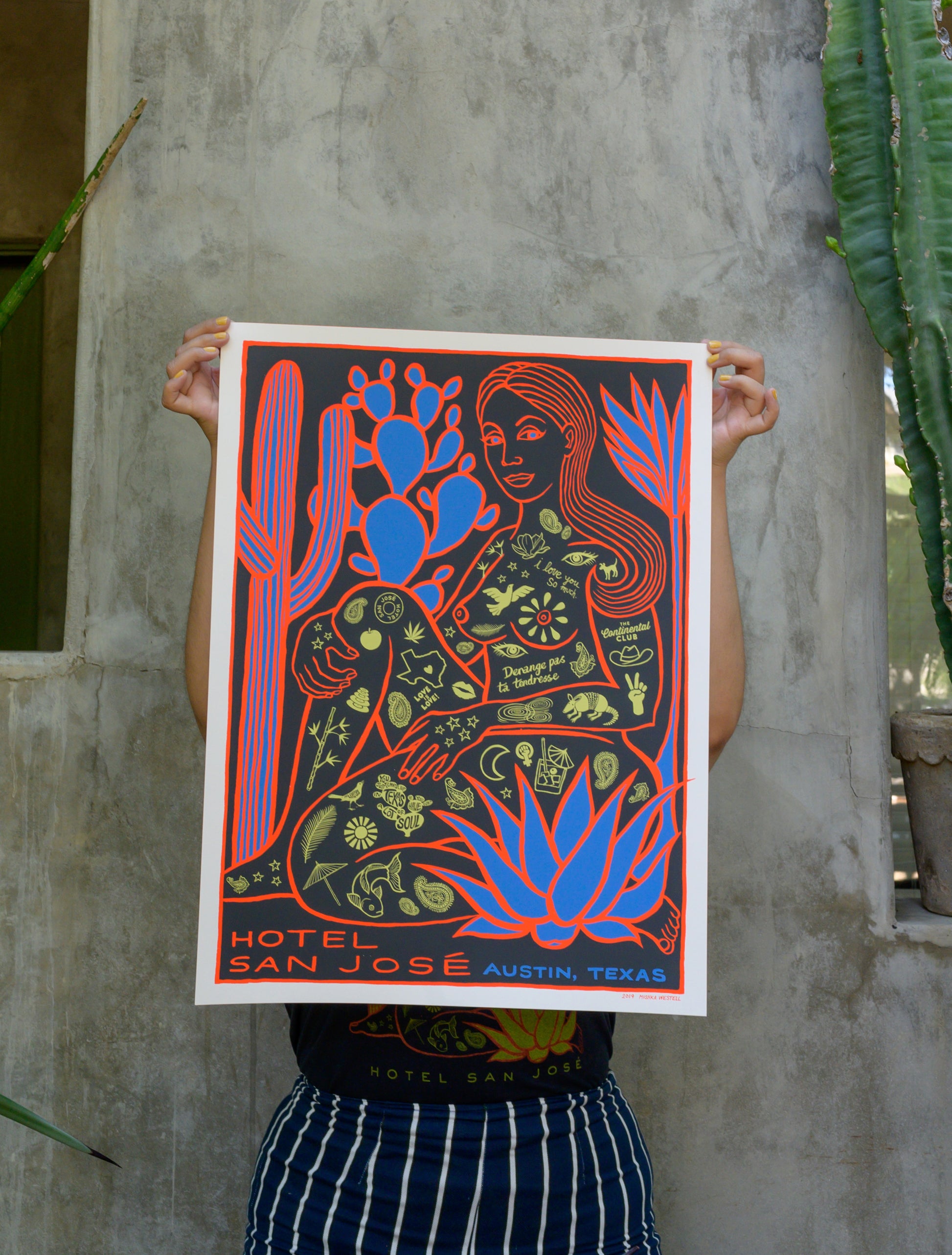 A poster featuring a stylized illustration of a woman covered in Austin's icon tattoos, sitting among cacti, with the text 'Hotel San Jose Austin, Texas' at the bottom being held by a person in front of cement wall with cacti plants on either side.