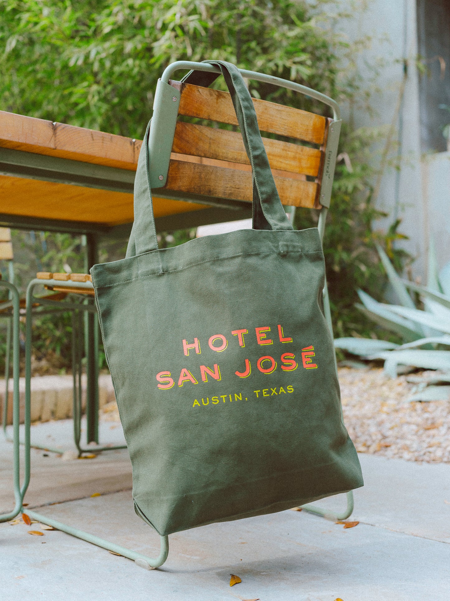 A green canvas tote bag with 'Hotel San Jose Austin, Texas' printed in orange and yellow lettering hanging on the side of a wooden chair with agave, bamboo and cement in the background.