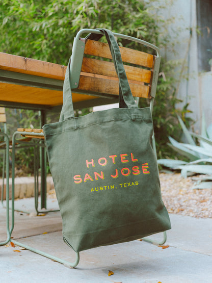 A green canvas tote bag with 'Hotel San Jose Austin, Texas' printed in orange and yellow lettering hanging on the side of a wooden chair with agave, bamboo and cement in the background.