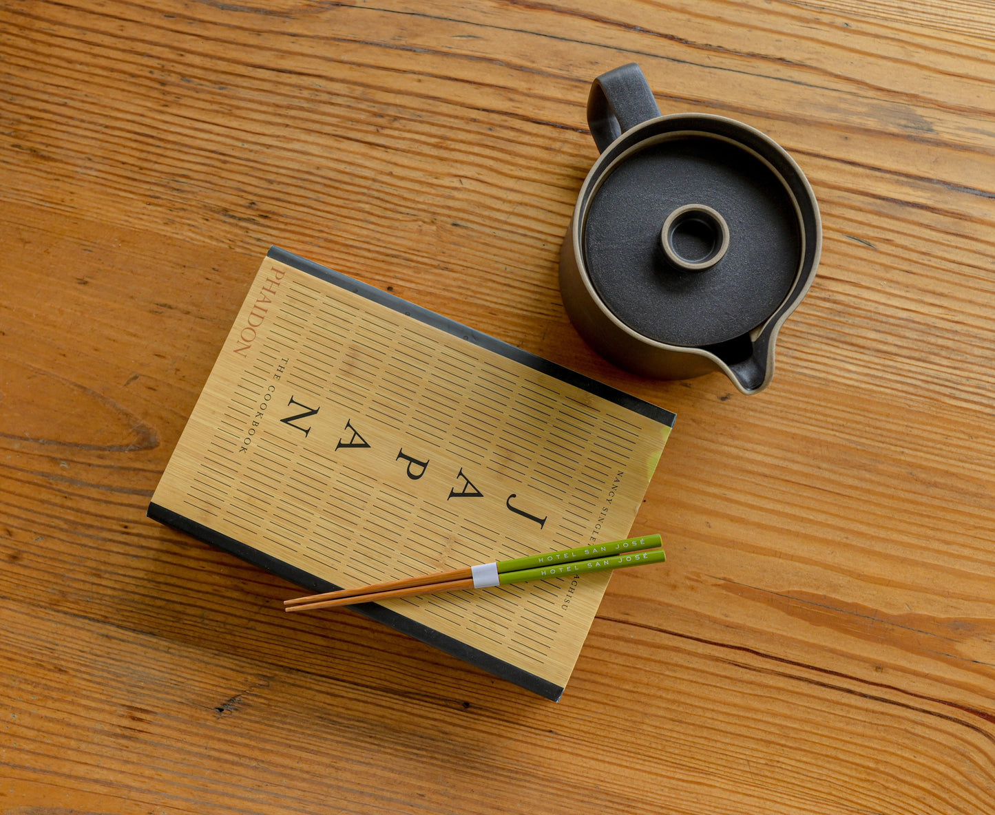Tea set with a teapot, book titled 'Japan', and 'HOTEL SAN JOSE' chopsticks on a wooden surface